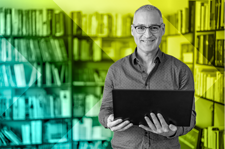 Smiling man holding a laptop in front of a bookshelf