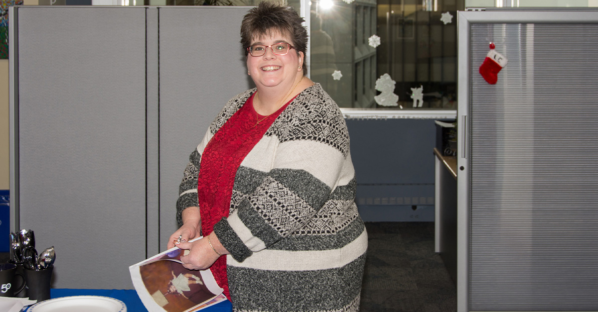 Lynn Blackburn stands in a CIA office cubicle holding a document, wearing a patterned knit sweater (2017).